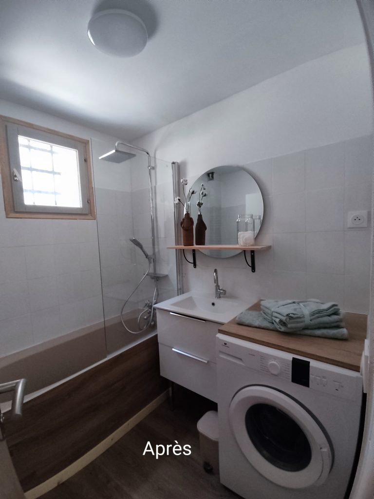 Salle de bain rénovée avec baignoire-douche, meuble vasque suspendu, miroir rond et machine à laver intégrée dans un espace optimisé et lumineux.